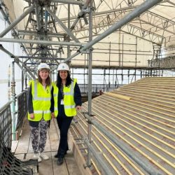 Two women in Hi Vis jackets and builder hats stood at the top of a building