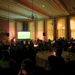 A dimly lit room with a keyboard player, violin player and a projection of a river behind them