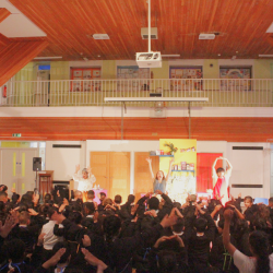 A group of primary school children dancing along with 3 performers on stage with their hands in the air