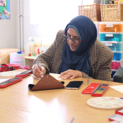 A photo of a elderly woman doing some watercolour painting. She is wearing a checkered coat and a blue hijab