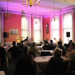 A band with a violin player, a bassist, a drummer and a pianist in a room with red and purple lights